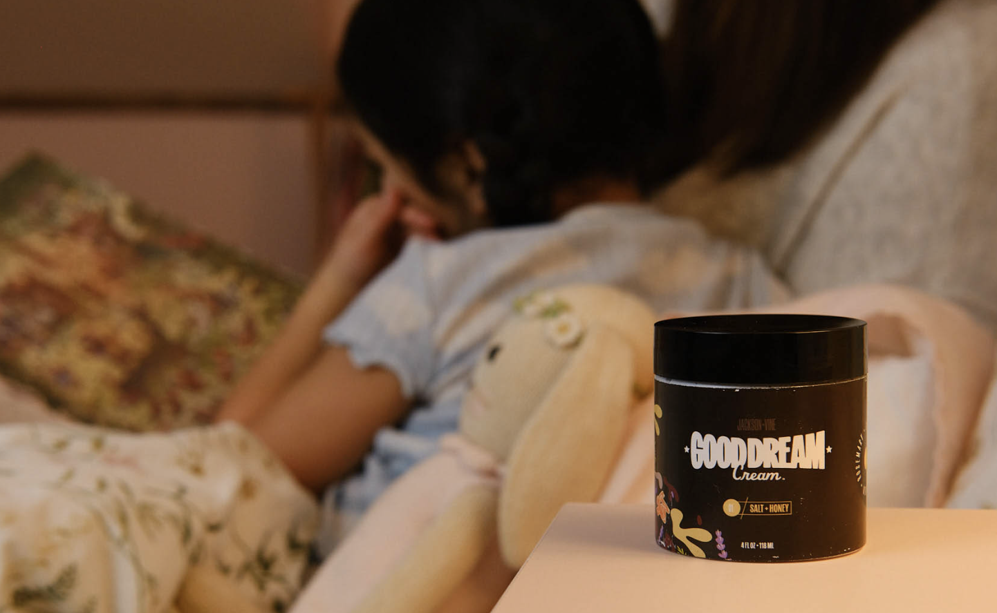 A jar of Good Dream Cream™ on a bedside table with a child resting in the background